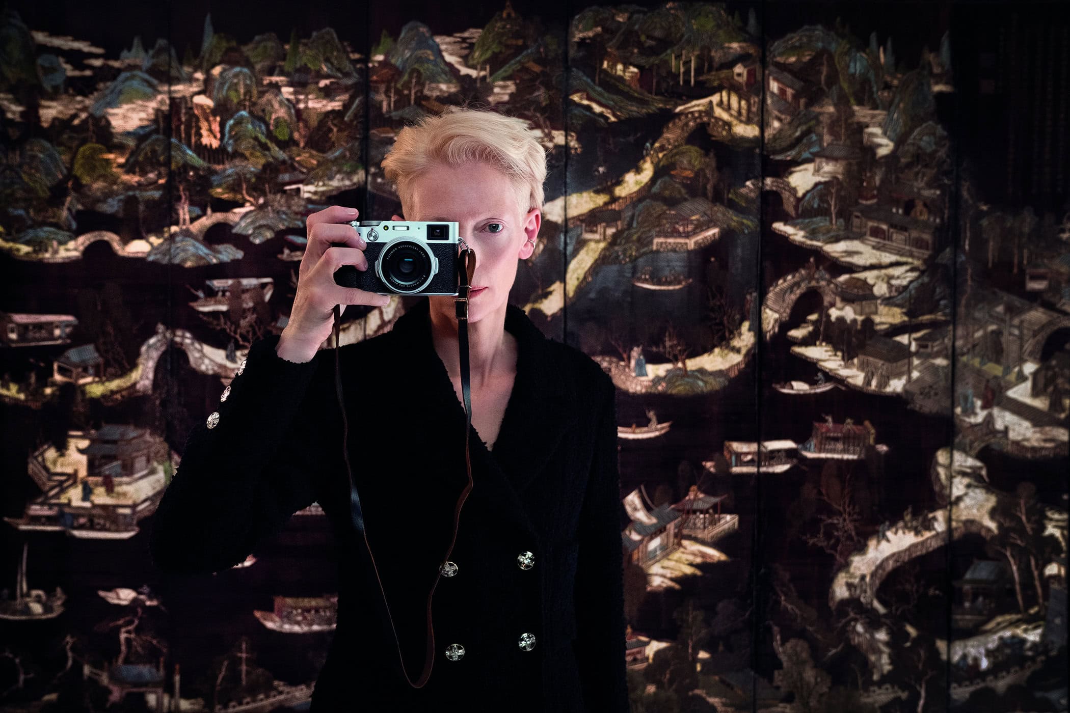 Tilda Swinton wearing a black double breasted jacket holding a vintage camera against an ornate patterned screen for Chanel
