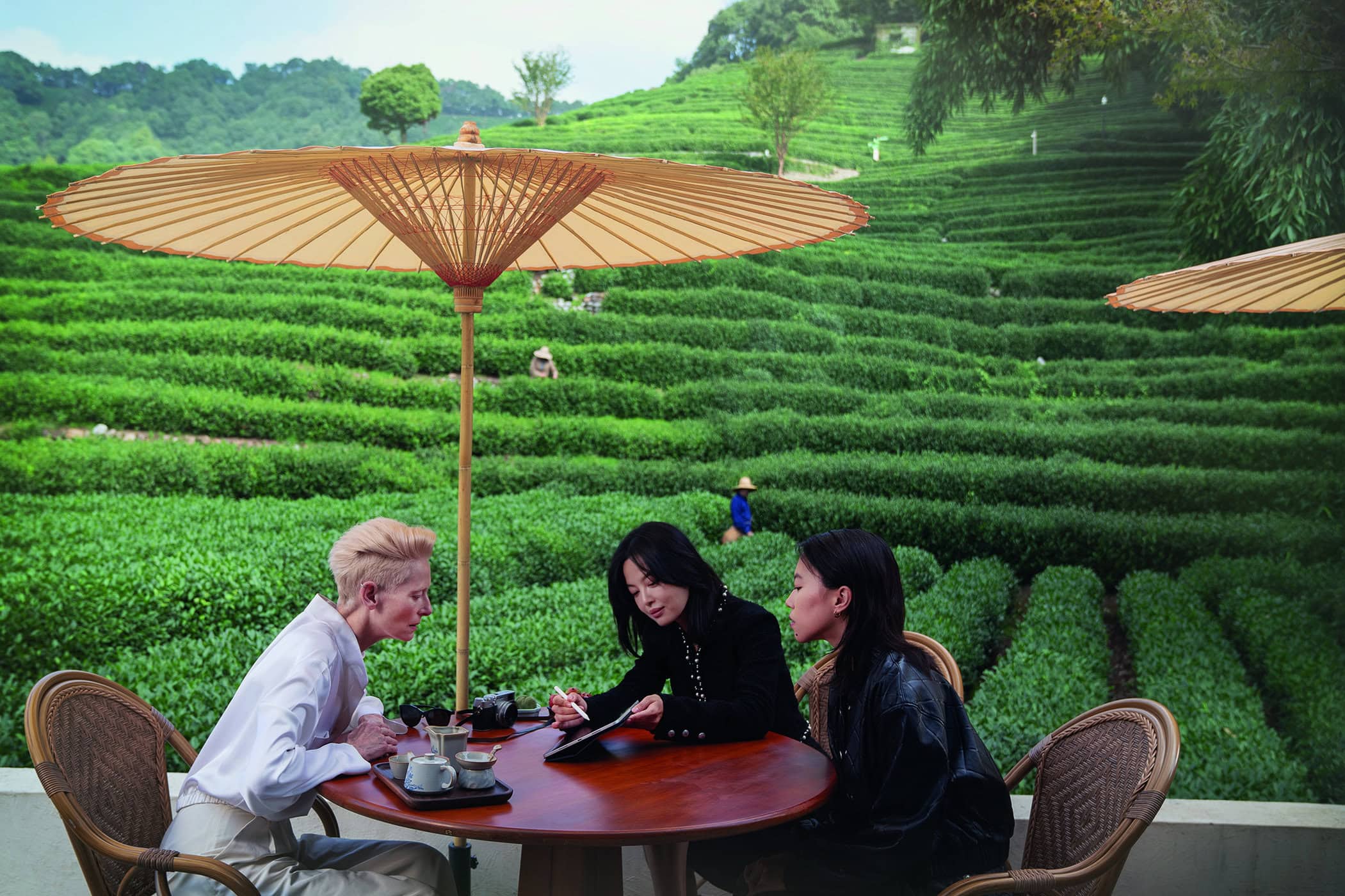 Three women seated at a round wooden table under a large beige paper parasol with lush terraced tea plantation hills behind them.