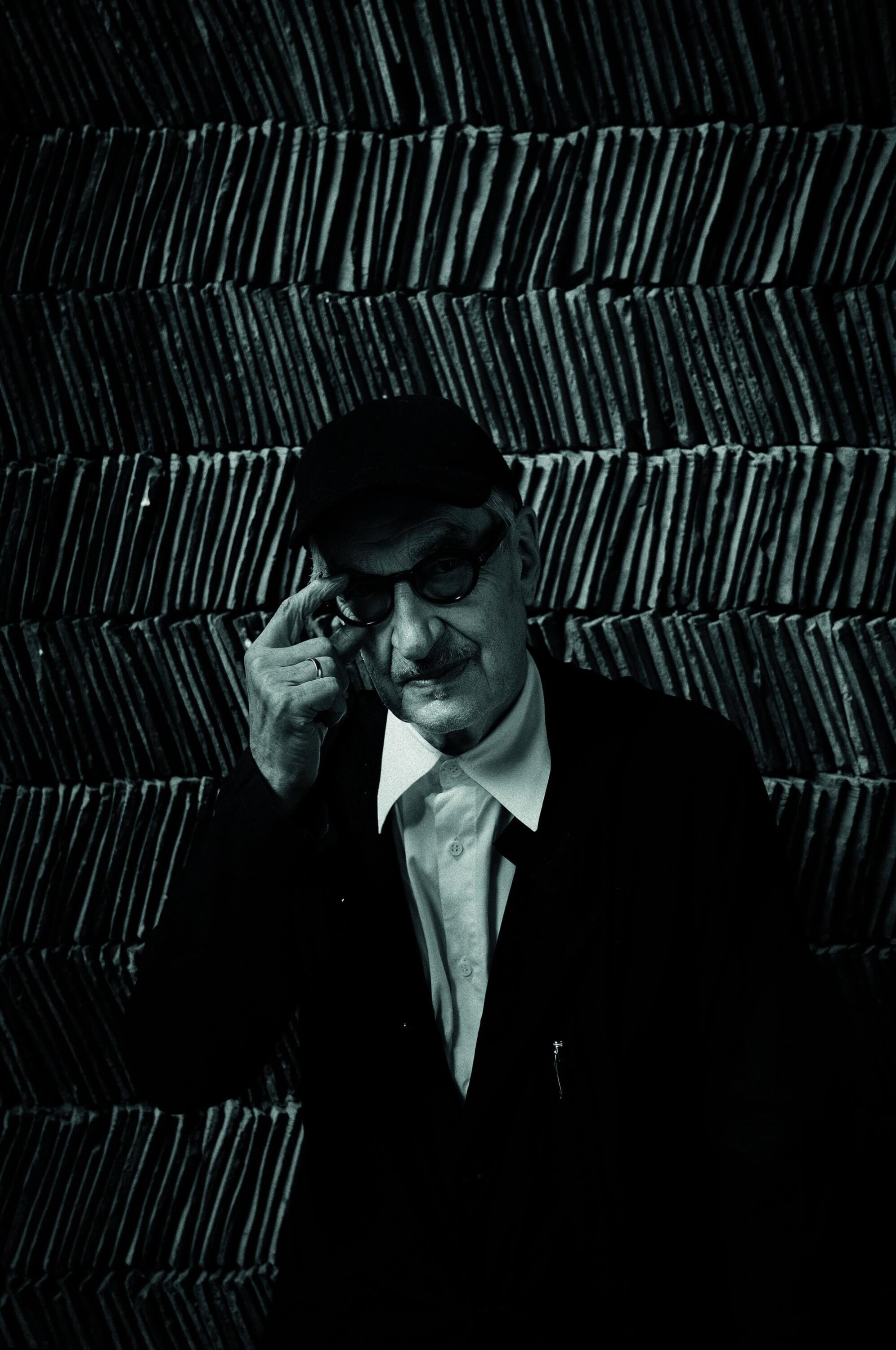 Man wearing a dark suit jacket, white shirt, cap, and glasses adjusting his eyewear against a textured vertical tile wall background