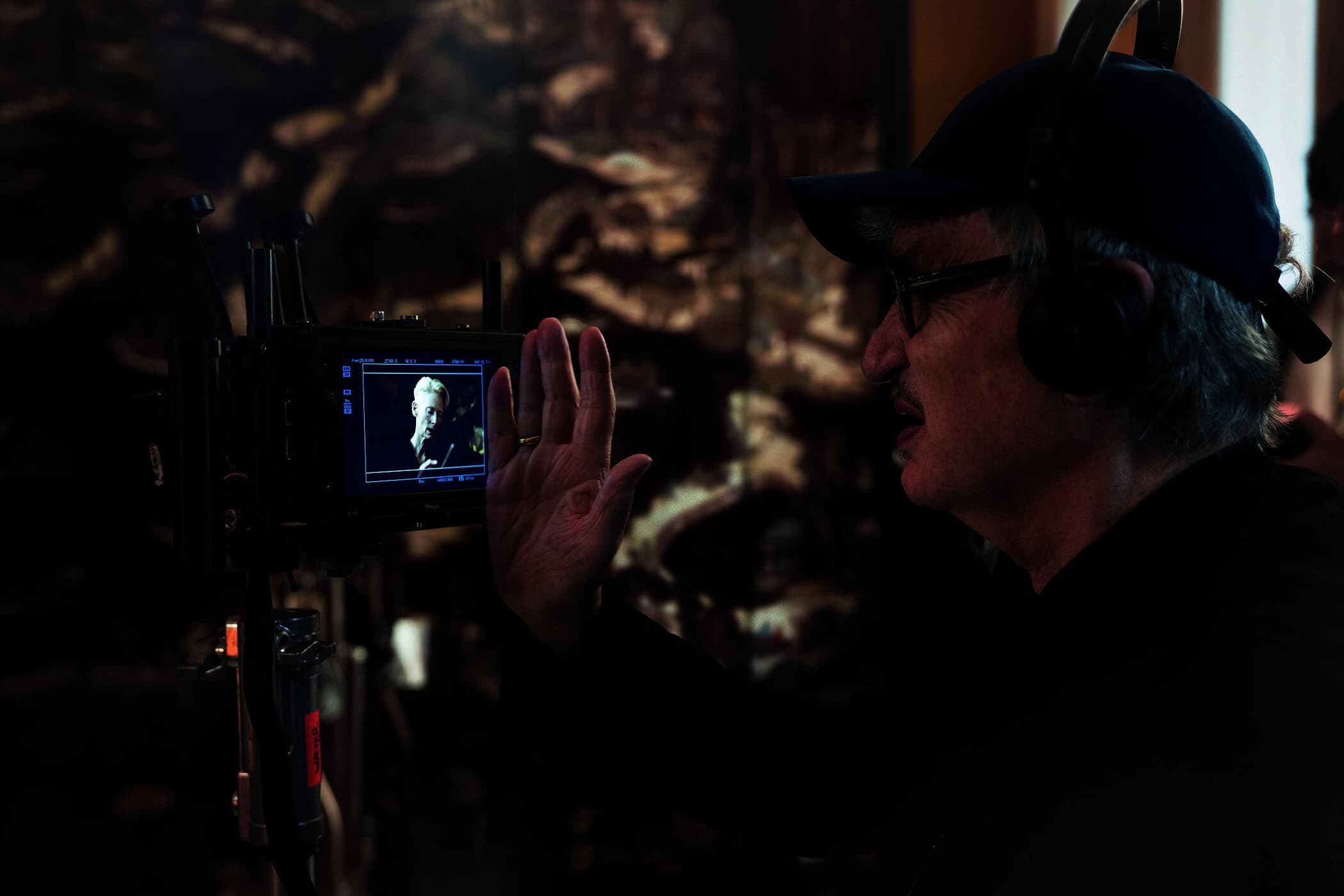 Wim Wenders wearing a cap and headphones on set, gesturing with a raised hand near a camera monitor showing Tilda Swinton.