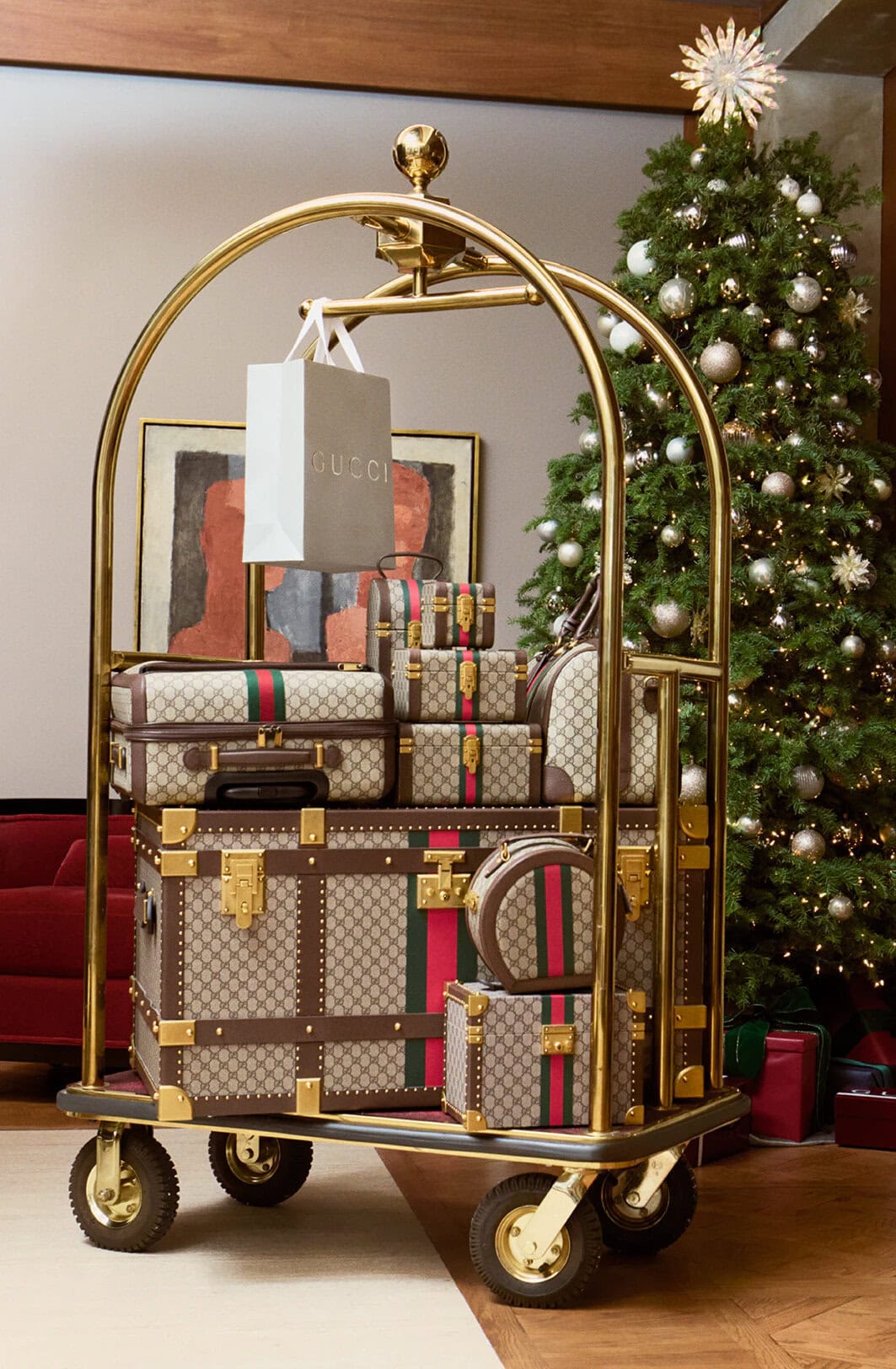 Gold luggage cart stacked with monogram luggage bags and a shopping bag beside a decorated Christmas tree