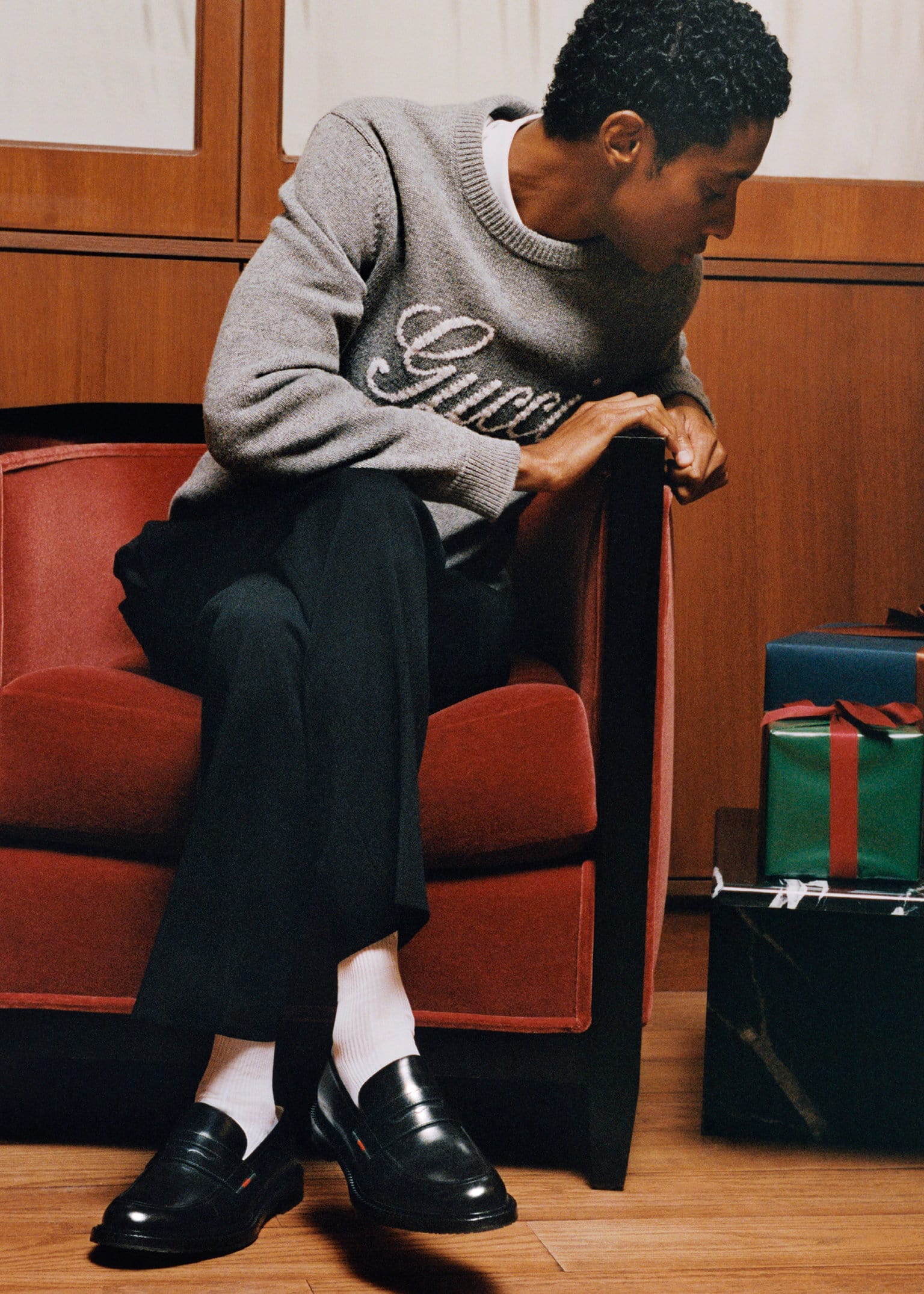 Man wearing grey Gucci sweater and black trousers seated in a red armchair next to wrapped gifts.