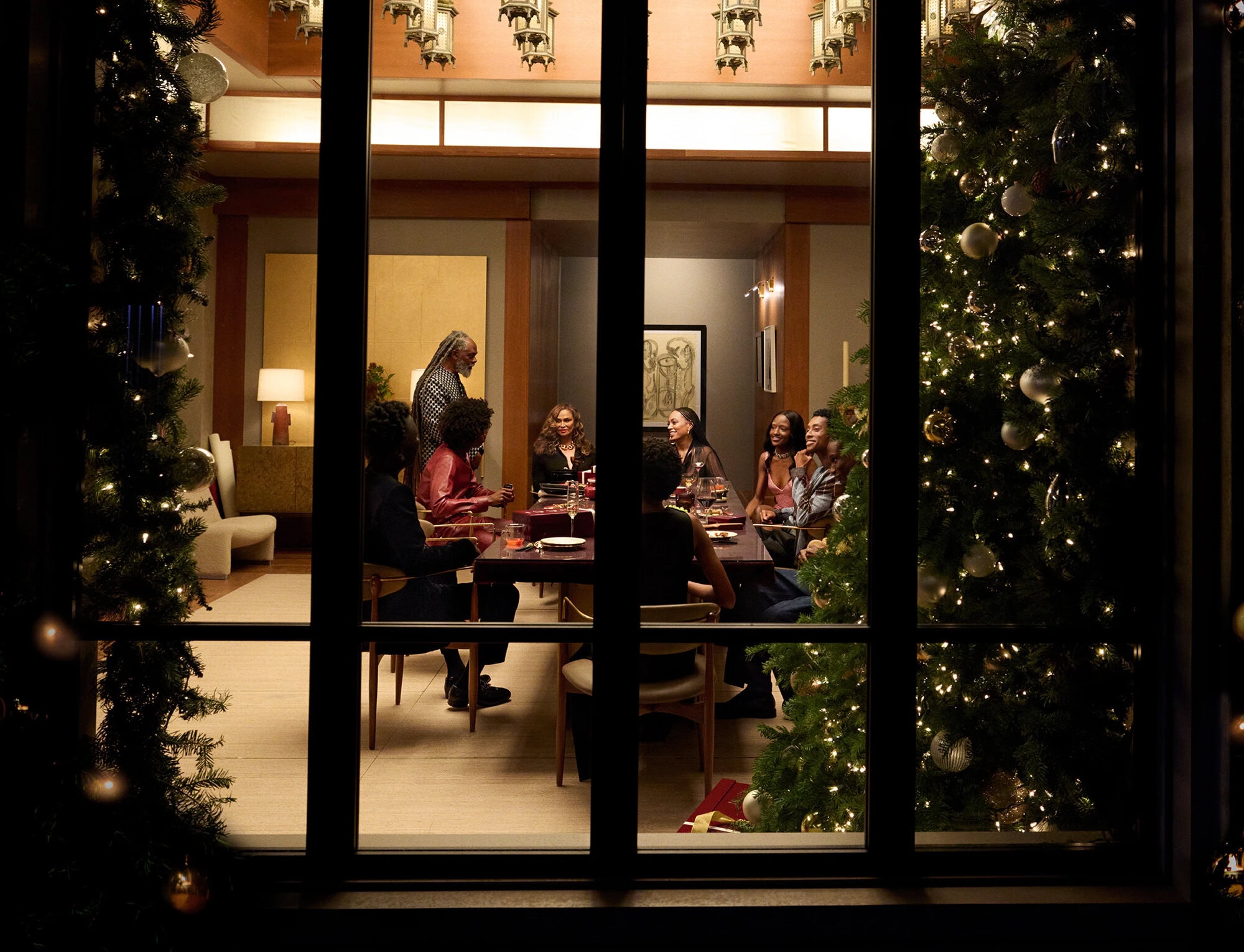 Group of people gathered around a dining table inside a warmly lit room, viewed through a large window framed by decorated Christmas trees.