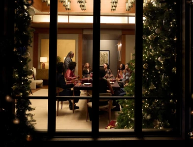 Group of people gathered around a dining table inside a warmly lit room, viewed through a large window framed by decorated Christmas trees.