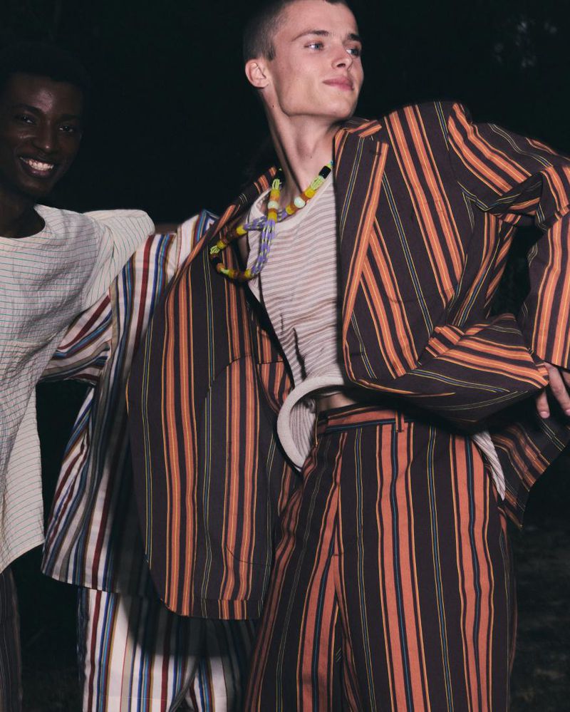 Two fashionable adult men in striped blazers and pants, one dark-skinned, one light-skinned, pose outdoors in soft lighting.