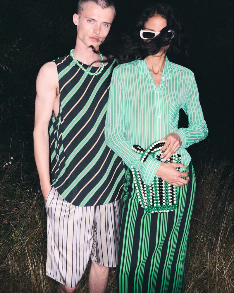 Two fashion models strike a pose outdoors, showcasing matching striped outfits. The man wears a tank and shorts, while the woman dons a blouse and skirt.