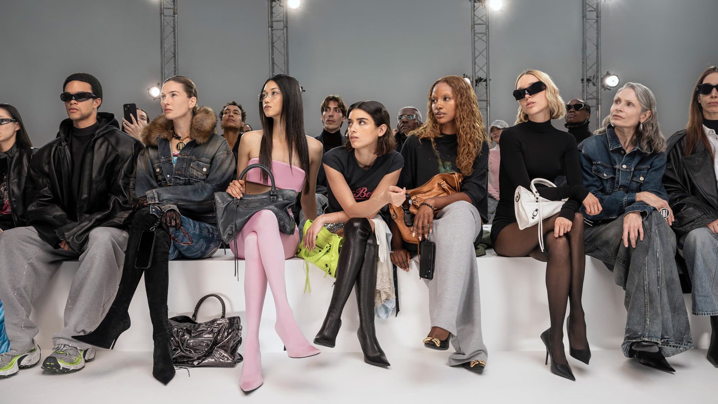 Group of models seated on a white bench wearing designer fashion and holding handbags for Balenciaga