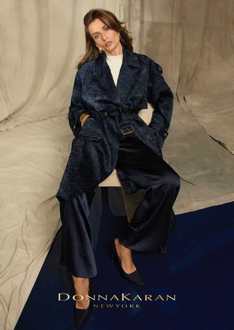 Model Andreea Diaconu seated in a navy blue coat and trousers against a draped neutral backdrop for Donna Karan.