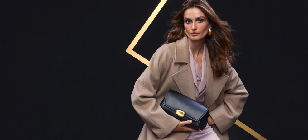 Model wearing a camel coat holding a black handbag against a dark background with golden lines