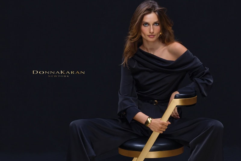 Model Andreea Diaconu in a black off-the-shoulder outfit seated on a modern chair against a dark background for Donna Karan.