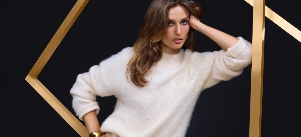 Model Andreea Diaconu wearing a fluffy white sweater leaning against gold geometric frames against a black background.