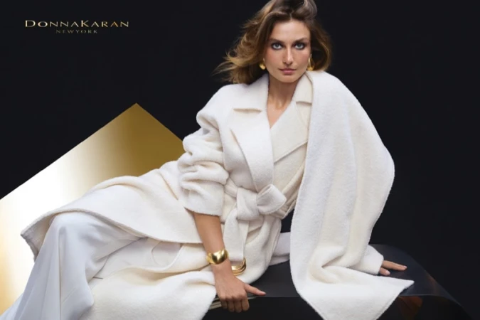 Model Andreea Diaconu wearing a cream belted coat and gold jewelry against a dark background for Donna Karan