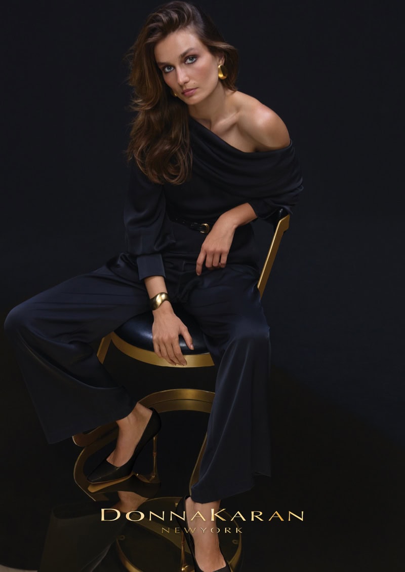 Model Andreea Diaconu seated on a modern chair wearing a black off-the-shoulder top and trousers against a dark background.