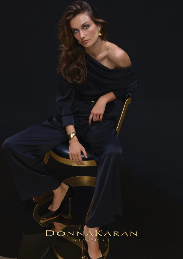 Model Andreea Diaconu seated on a modern chair wearing a black off-the-shoulder top and trousers against a dark background.
