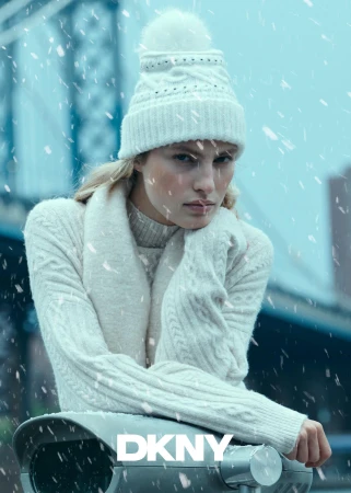 Model in white cable knit sweater and matching beanie leaning on a railing in falling snow