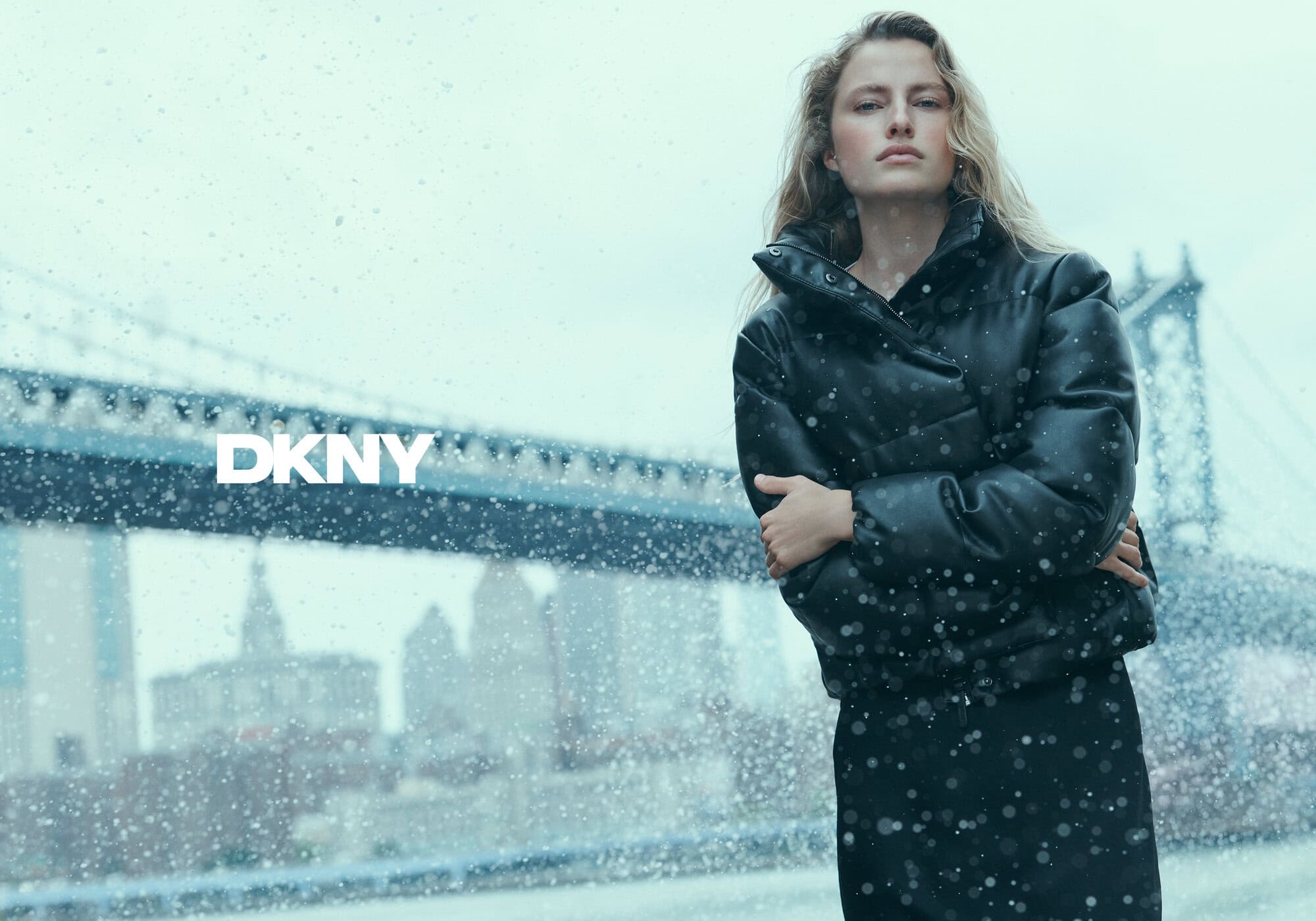 Model wearing a black puffer jacket against a bridge backdrop during winter snow for DKNY