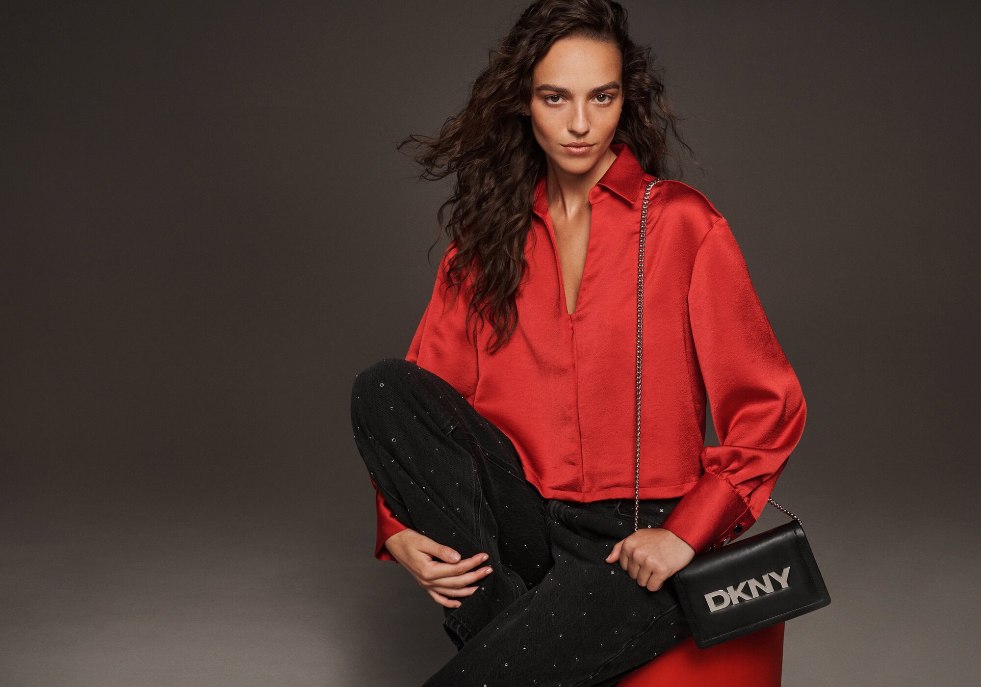 Model wearing a vibrant red satin blouse and dark patterned trousers holding a black DKNY chain shoulder bag in a studio setting.