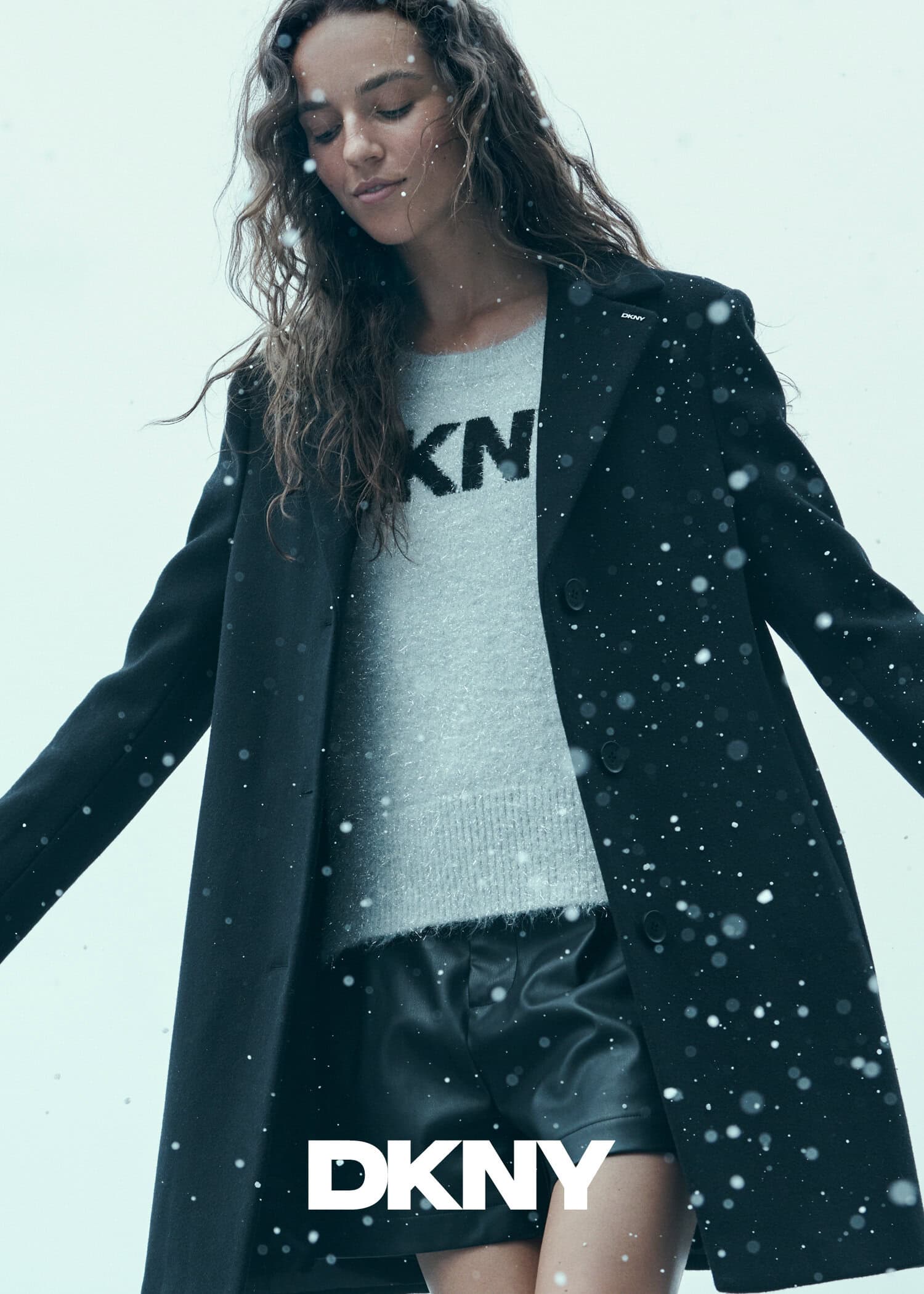Model wearing a black coat and grey sweater with snowflakes falling against a pale sky background