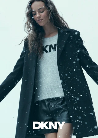 Model wearing a black coat and grey sweater with snowflakes falling against a pale sky background