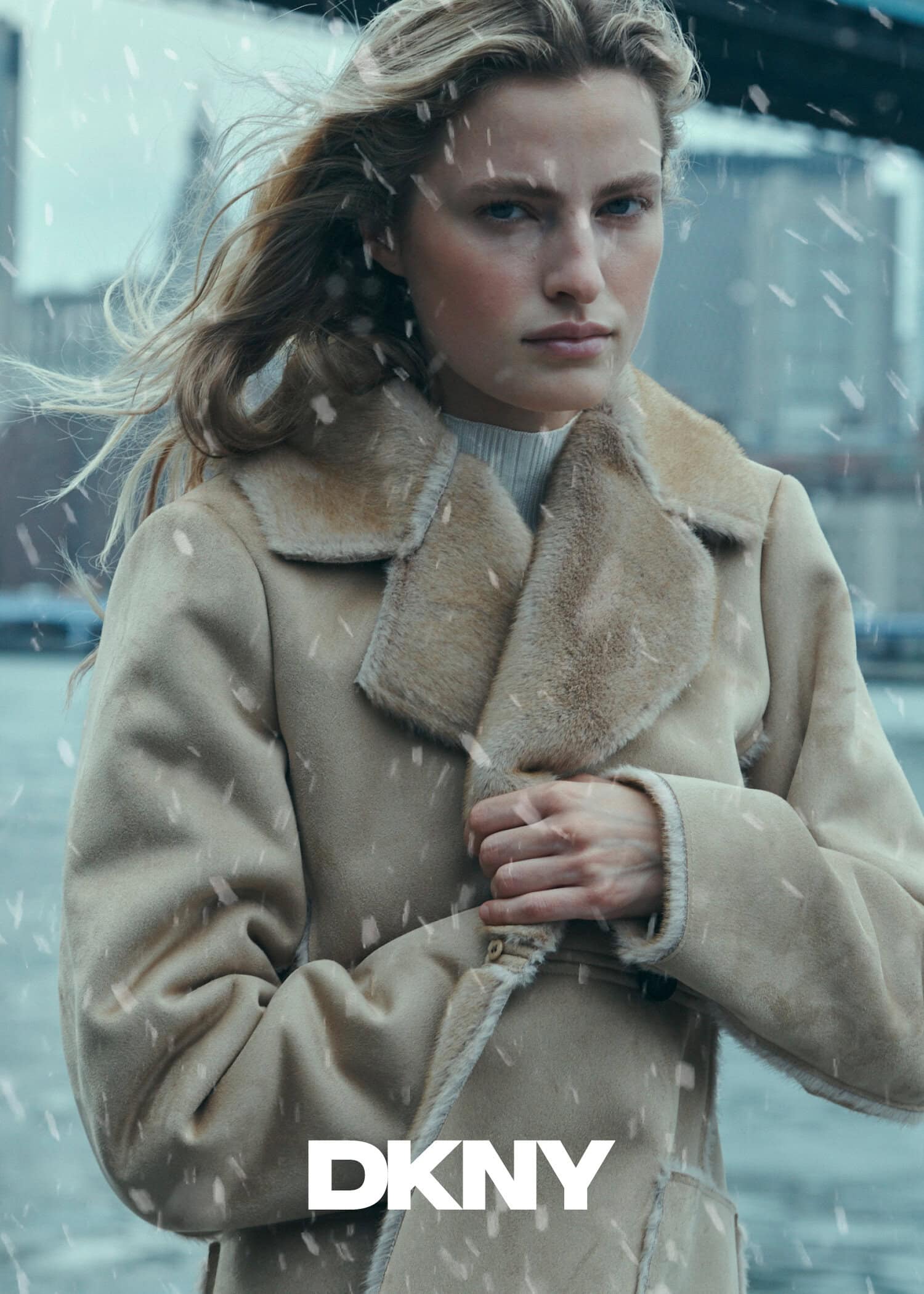 Model in a tan shearling coat outdoors in the snow with a bridge in the background