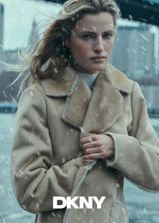 Model in a tan shearling coat outdoors in the snow with a bridge in the background