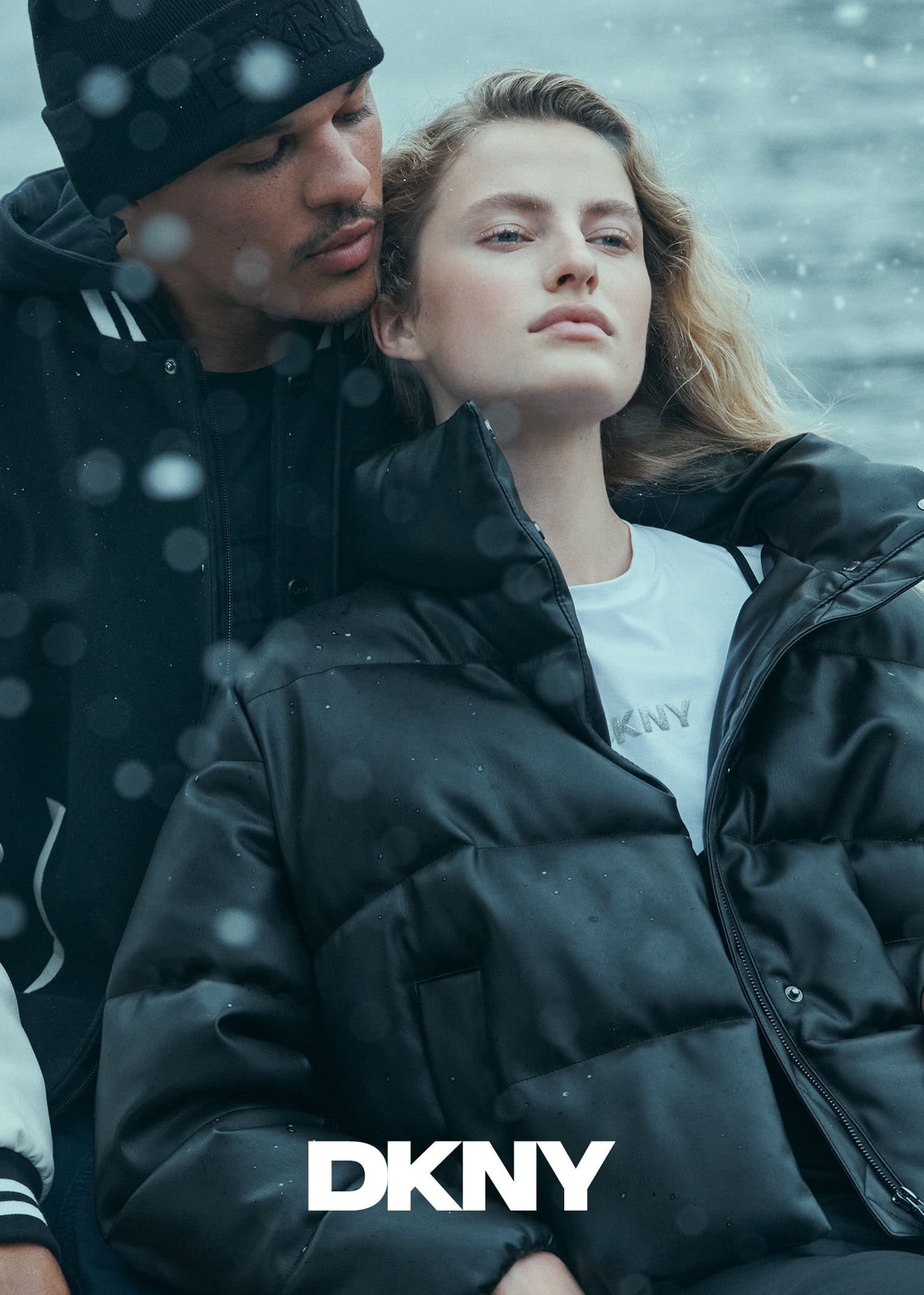 Man in black beanie and jacket embracing a woman in a black puffer coat with DKNY branding outdoors.