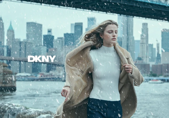 Model in a cream shearling coat and white turtleneck against a snowy waterfront skyline for DKNY.