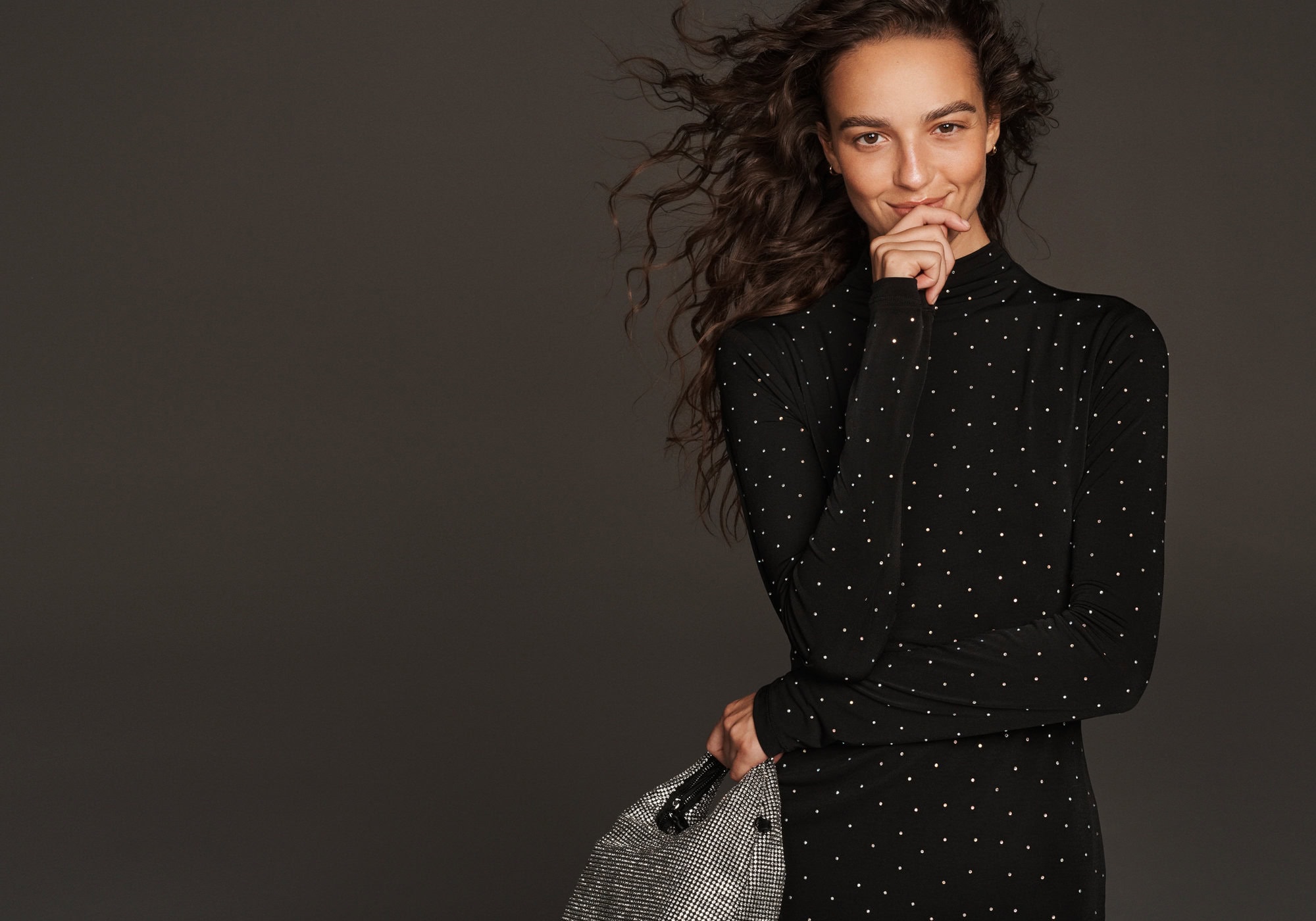 Woman in a black dress with white dots holding a metallic handbag against a dark grey background