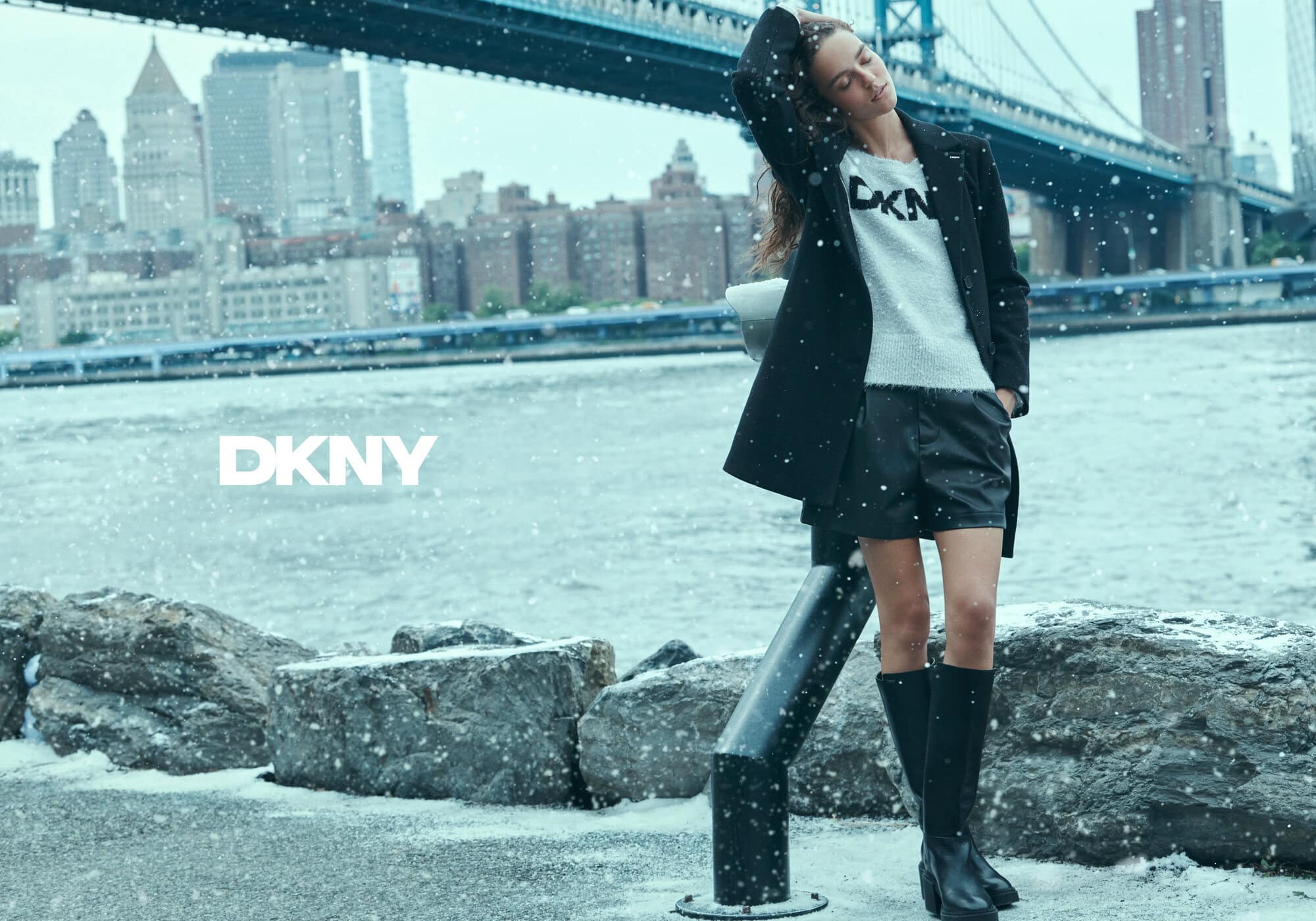Model wearing a black coat and shorts standing near a river with a bridge and city skyline under falling snow.