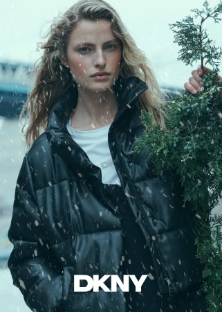 Model in a black puffer jacket holding evergreen branches amidst falling snow for the DKNY Holiday 2024 campaign
