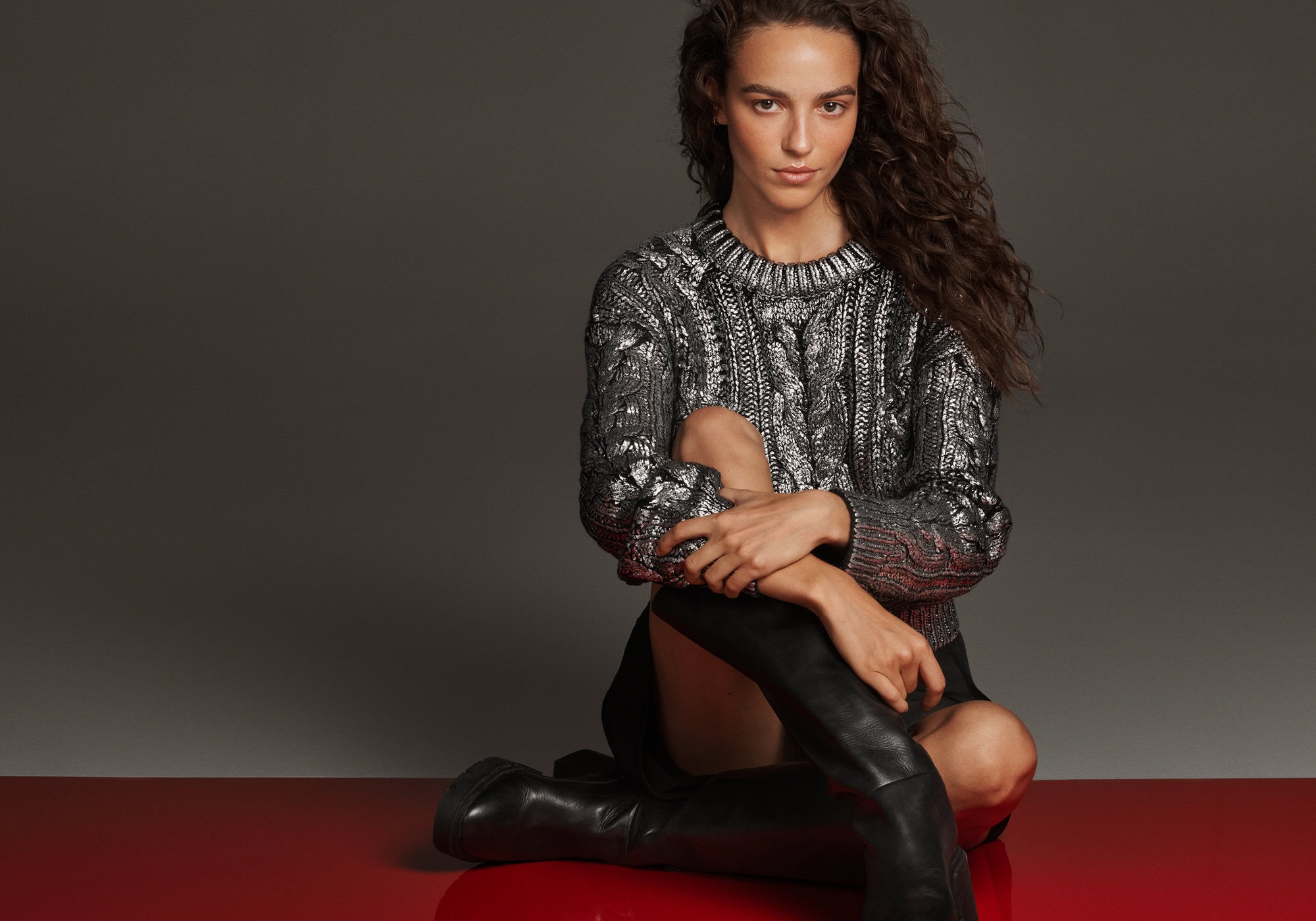 Model seated on a reflective red surface wearing a metallic grey sweater and black boots against a dark grey studio background.