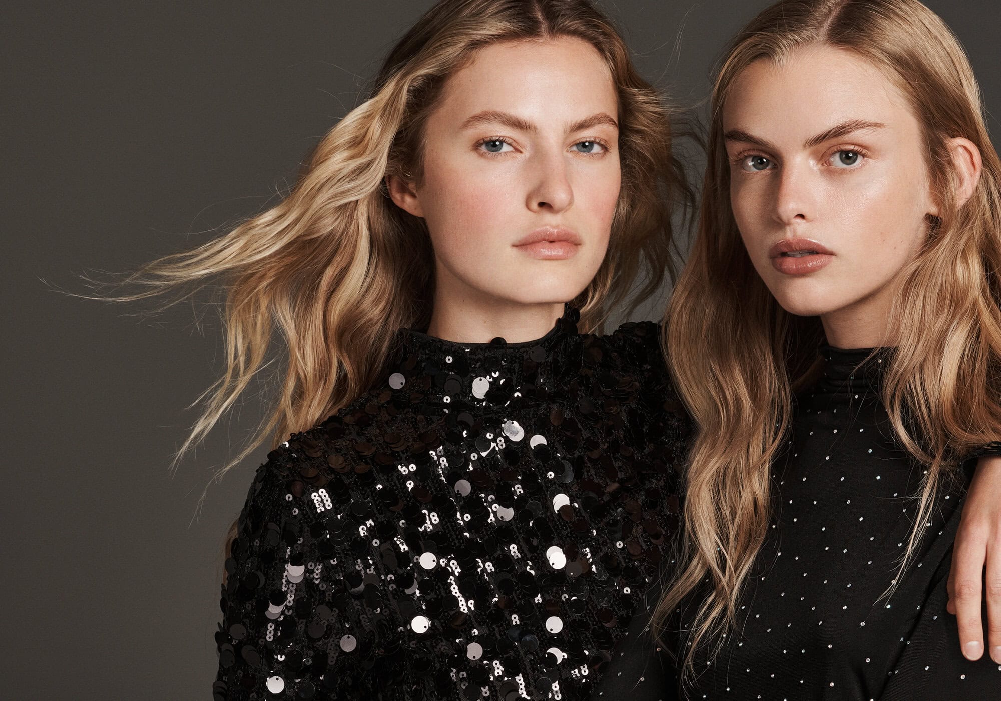 Two female models with blonde hair wearing black sequined tops against a plain grey studio background.