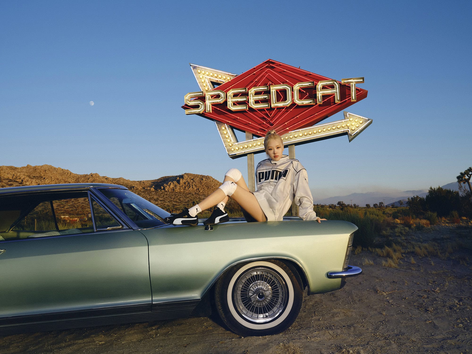 Rosé Park sitting on the hood of a vintage light green car wearing a Puma windbreaker and sneakers beneath a neon Speedcat sign.