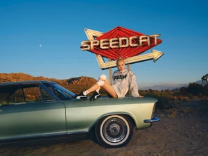 Rosé Park sitting on the hood of a vintage light green car wearing a Puma windbreaker and sneakers beneath a neon Speedcat sign.