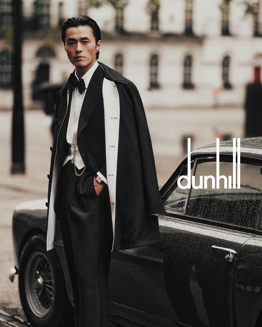 Man wearing a tuxedo and dark coat leaning against a vintage car on a street