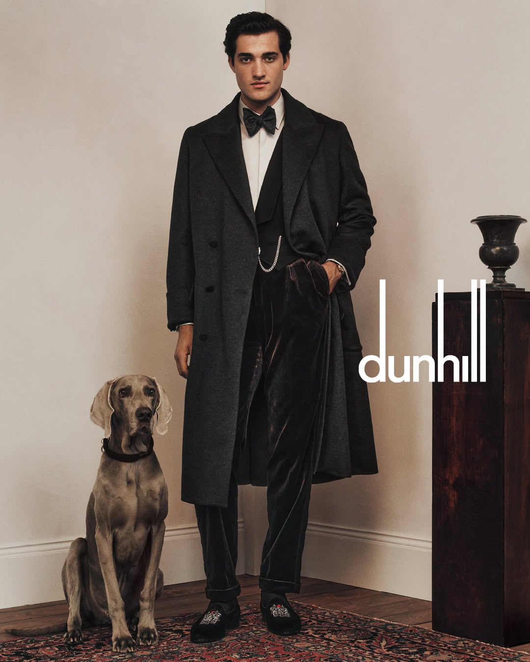 Man in dark overcoat and velvet trousers standing next to a dog on an ornate rug for Dunhill
