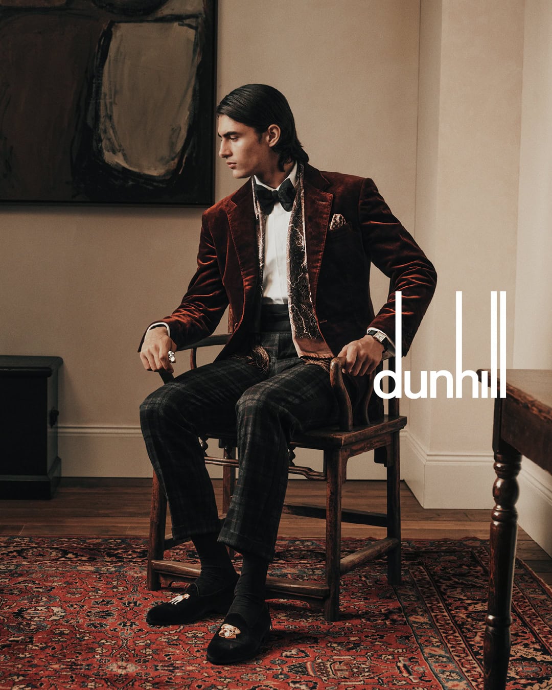 A man wearing a velvet blazer and patterned trousers sitting on a wooden chair in an interior room.