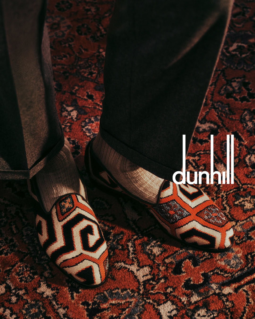 Close-up of patterned loafers and dark trousers standing on a patterned oriental rug with the dunhill logo visible