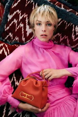 Model Edie Campbell in a vibrant pink blouse holding an orange leather handbag with gold hardware against a patterned red cushion background.