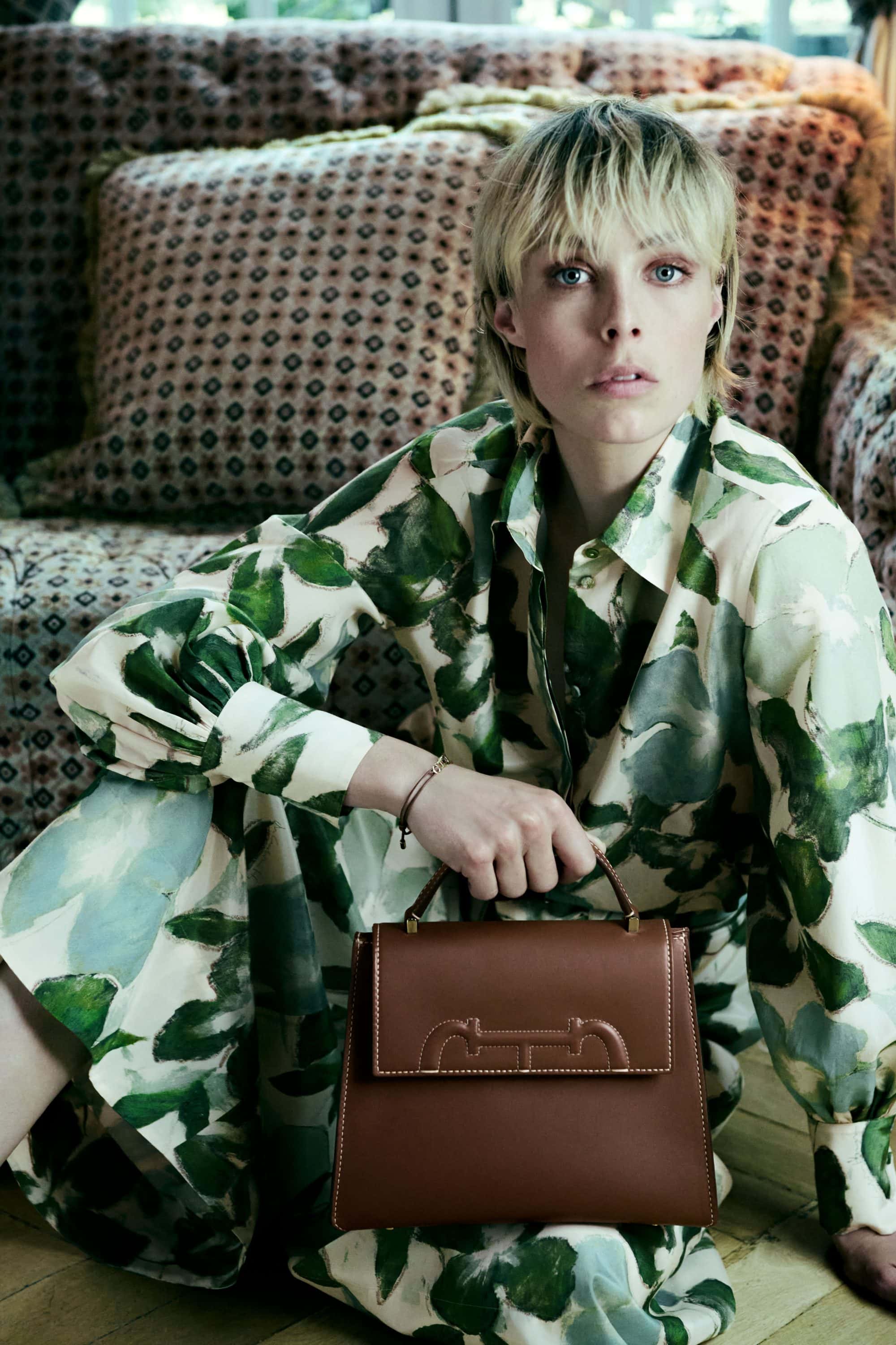 Model Edie Campbell holding a brown leather handbag, wearing a green patterned dress against a tufted sofa for CH Carolina Herrera.