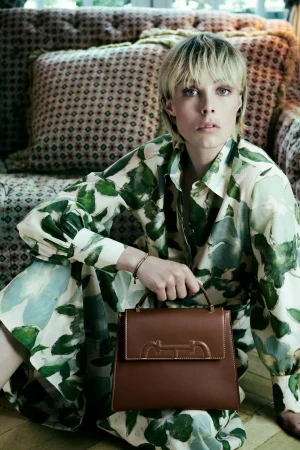 Model Edie Campbell holding a brown leather handbag, wearing a green patterned dress against a tufted sofa for CH Carolina Herrera.