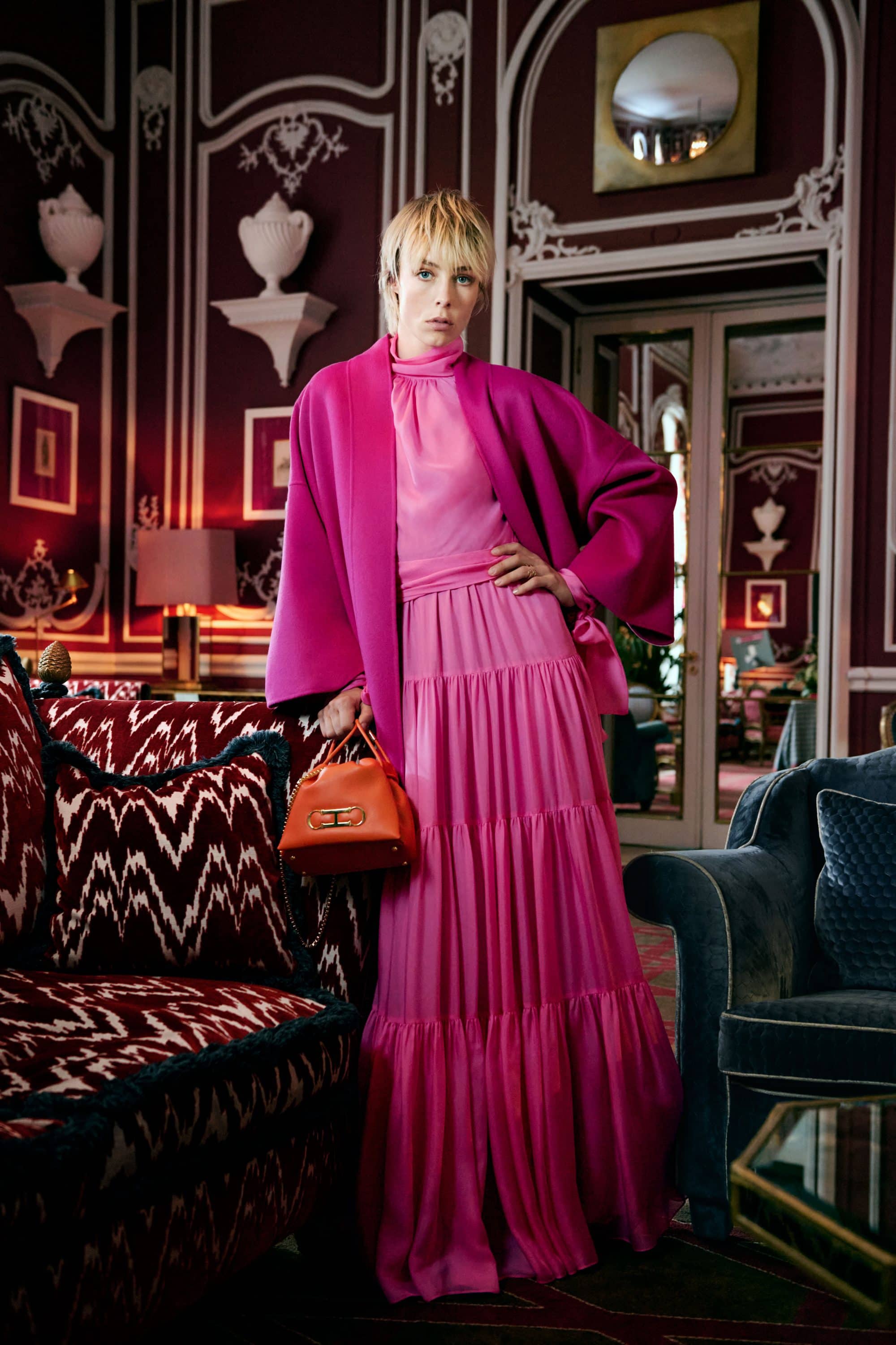 Model Edie Campbell in a pink tiered dress and coat holding an orange handbag inside a hotel lobby interior
