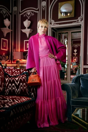 Model Edie Campbell in a pink tiered dress and coat holding an orange handbag inside a hotel lobby interior
