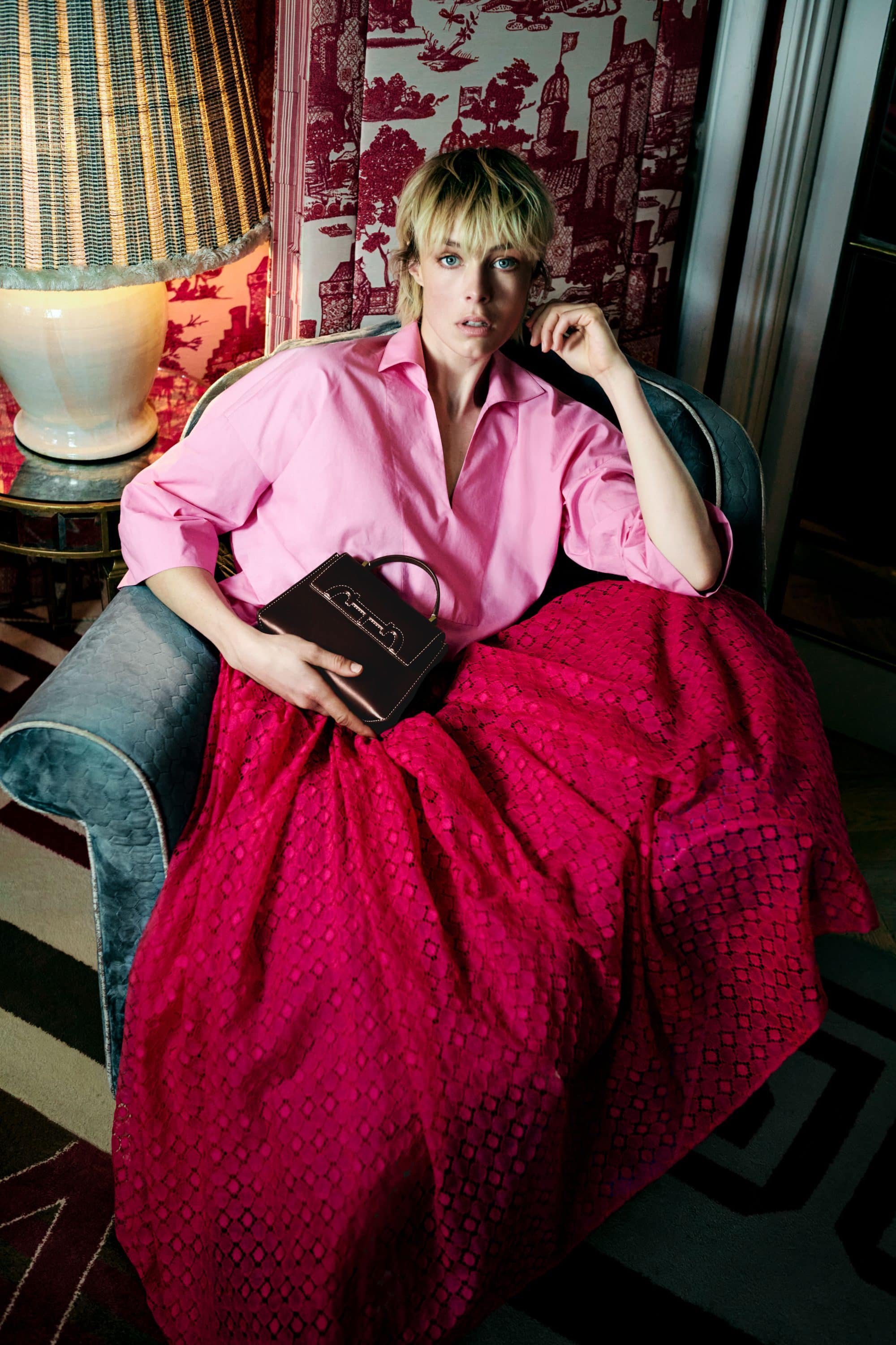 Model Edie Campbell seated in a green armchair wearing a pink shirt and red skirt holding a dark handbag.