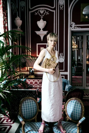 Model Edie Campbell in a white dress holding a woven handbag inside an ornate room