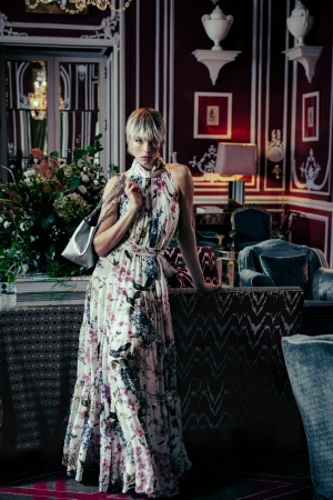 Model Edie Campbell in a floral print halter dress holding a silver handbag in a richly decorated room