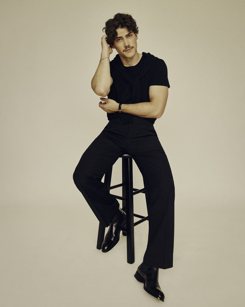 Portrait of a young adult male model with dark hair and a mustache, wearing black clothing and sitting on a stool against a neutral backdrop.