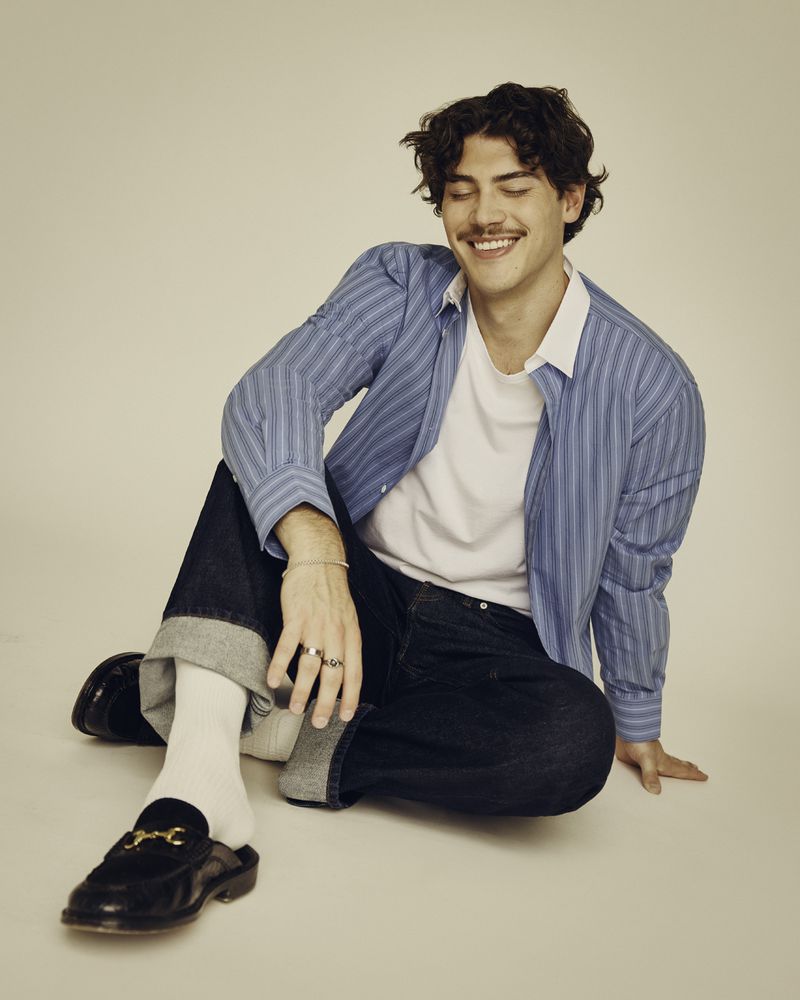 Smiling man in a blue striped shirt sits indoors on the floor, with cuffed jeans, white socks and black leather loafers with gold buckles.