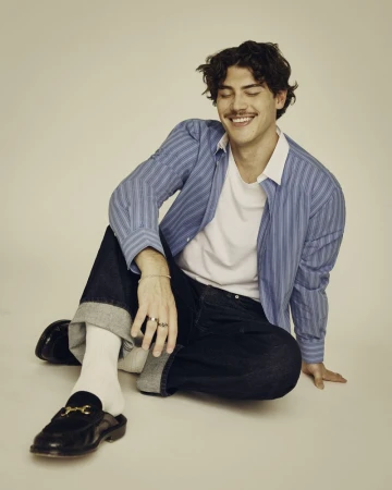 Smiling man in a blue striped shirt sits indoors on the floor, with cuffed jeans, white socks and black leather loafers with gold buckles.