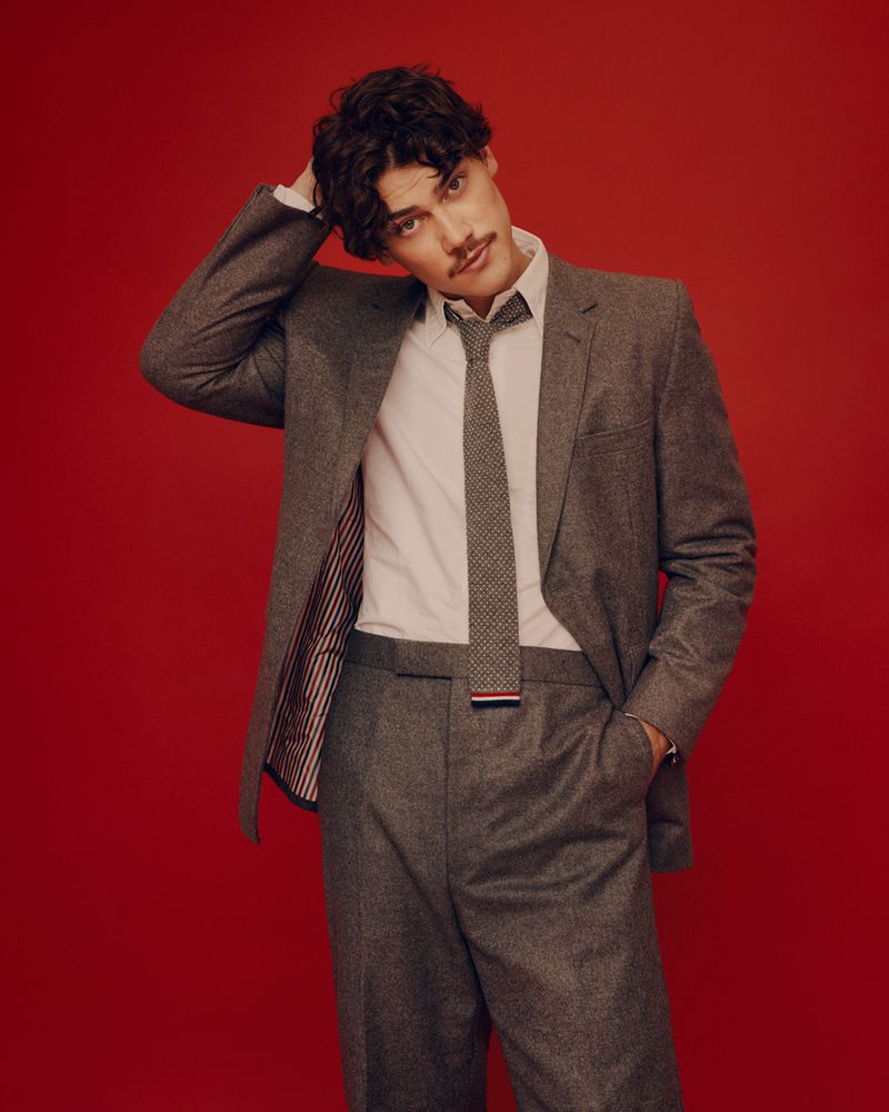 Adult male in a grey suit and tie poses against a red backdrop, his hand resting on his head in a fashionable photo studio portrait.